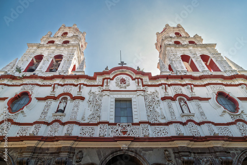 Low angle view of ornate white and red Baroque facade of Expiatory Temple of the Holy Spirit with twin bell towers and Jesuit IHS symbol against clear blue sky in Puebla, Mexico