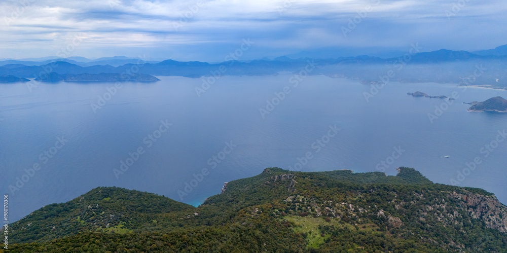 Obraz premium Sweeping aerial drone landscape looking out across a magnificent coastal bay featuring deep blue waters framed by rolling green forested mountains on a serene cloudy day