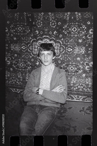 Vintage black and white portrait of young man. Retro 35mm film scan of young guy sitting in front of patterned wall carpet. Authentic USSR or Eastern Europe 80s aesthetic, nostalgia or retro fashion