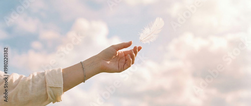 Close view of a human hand reaching toward a floating white feather against a soft cloud background, lit by warm natural light, suggesting gentleness, release and quiet serenity in still air