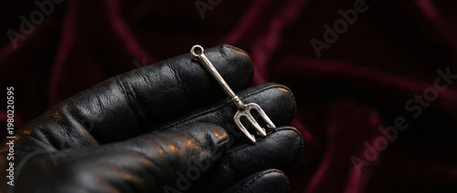 Close up of a small silver trident charm resting on a black leather gloved hand against a dark red velvet background, emphasizing symbolism, mystery, danger, ornament detail, and contrast