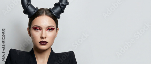 Studio beauty portrait of a woman with sculptural black horn headpiece, dramatic red winged eyeliner and dark lipstick, styled in a sharp black jacket against a clean gray background