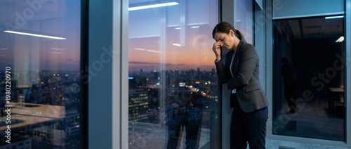 Stressed businesswoman in a gray suit stands by a floor to ceiling window at dusk, looking down with hand to her forehead as city lights emerge beyond the quiet modern office after a long day