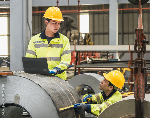 Two professional engineer safety vest hard hat collaborating in factory use laptop measuring tape inspect steel coil representing industrial teamwork quality control manufacturing excellence warehouse