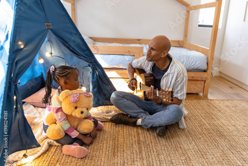 African American father and child sitting on bedroom rug with teepee, playing guitar, hugging teddy