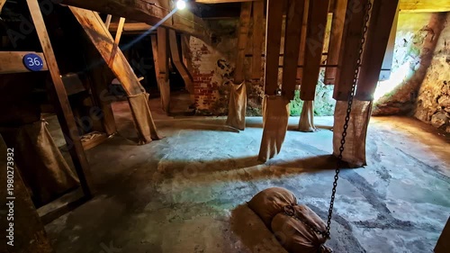 Old mill interior with hanging fabric chutes and a chained sandbag counterweight on a dusty floor. Rustic industrial heritage scene with moody light and texture.