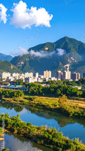 A vibrant landscape showcases a tranquil river winding through lush greenery, framed by a backdrop of towering mountains and a modern urban settlement under a clear blue sky.