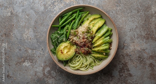 Top down composition of a plant based bowl including avocado, green beans, zucchini noodles, with green dressing on a rustic concrete backdrop