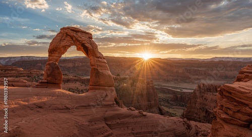 Warm sunrise light brightens a prominent rock arch within an expansive canyon scene with clear and cloudy sky