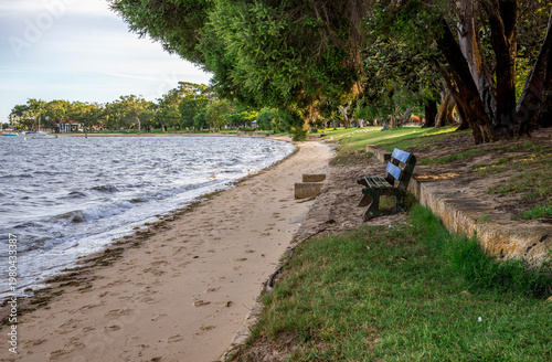 Sandy beach with wooden bench beside Swan River under trees in Matilda Bay Reserve, Perth, Australia, 2 February 2022