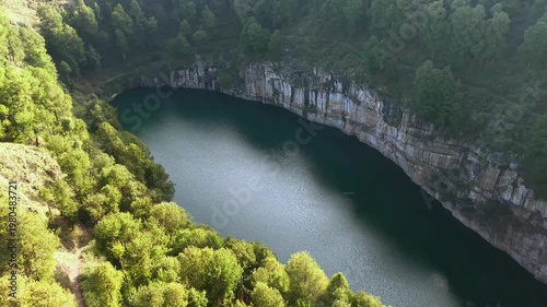 Aerial view reveals a deep, calm crater lake surrounded by lush green trees. Rocky cliffs encircle the dark water, creating a serene natural amphitheater. Shadows stretch across the water