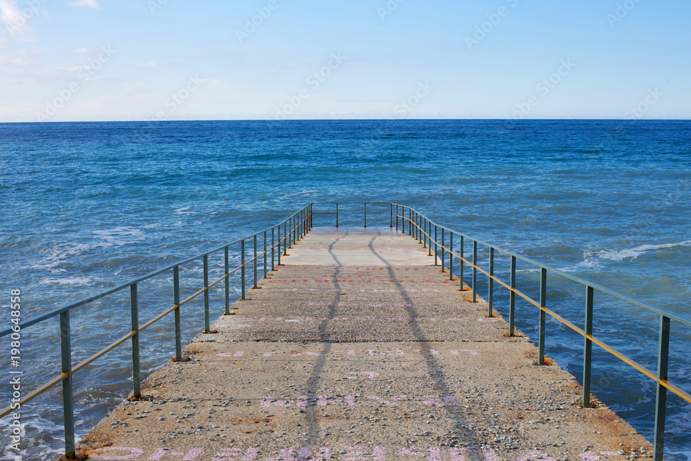 Fototapeta premium Pier leading into blue sea with horizon, minimal seascape. Concrete jetty with railings, calm water and clear sky, symbol of travel, freedom, infinity and peaceful summer vacation