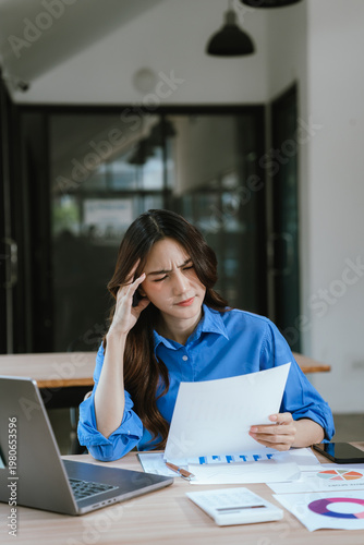 A businesswoman looks stressed while reviewing financial reports beside a laptop, analyzing data and facing challenges in a modern office workspace.