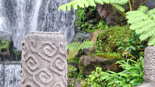 Schilderij op canvas Ancient Engraved Stone Pillar Beside Cascading Tropical Waterfall in Lush Fern G