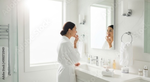 A woman in a white robe applying lipstick in front of a mirror in a bright, well-lit bathroom with a white marble countertop.