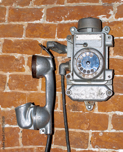 Old rotary phone mounted on orange brick wall with a receiver hanging down