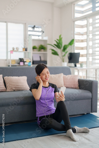 Asian adult woman sportswear enjoy listening music phone app sitting yoga mat home living room modern bright apartment fitness relaxation wellbeing wellness happy lifestyle