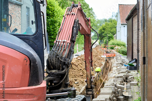 An excavator digs a long trench for utility installation alongside a residential road. Paving stones are removed, and a worker is visible in the ditch, indicating ongoing urban construction work.