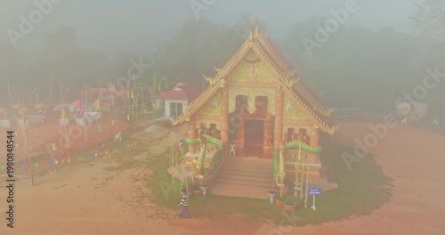 Aerial view of a beautiful golden Buddhist temple in Sapan Village, Nan surrounded by lush forest and morning mist. The tranquil rural landscape and sea of fog create a spiritual atmosphere