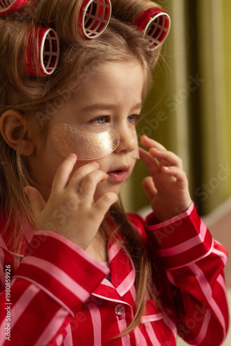 Cute little girl wearing hair rollers and applying under eye patches at home. Concept of skincare, self-care routine, childhood lifestyle, and beauty habits. Warm natural light and cozy atmosphere