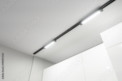 Foto A modern interior with a sleek black track lighting system mounted on a white ceiling, illuminating clean white cabinetry below