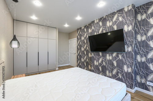 A modern bedroom with a large bed, patterned dark wallpaper, wall-mounted TV, built-in wardrobe, pendant light, recessed ceiling lights, wooden flooring, neutral tones, minimalist design.