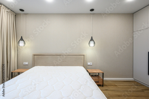 A minimalist bedroom with a large bed, beige upholstered headboard, two wooden bedside tables, pendant lights, neutral walls, wooden flooring, light curtains, built-in wardrobe.