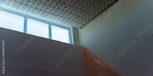 The image captures the inside of a vacant room with a window, a vintage patterned ceiling, and walls in the process of renovation, symbolizing a home improvement project