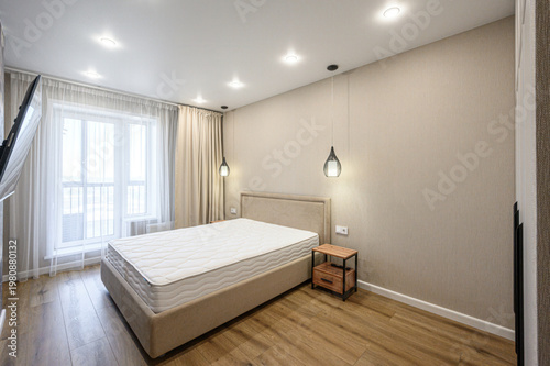 A minimalist bedroom with a large bed, beige upholstered headboard, wooden nightstand, pendant lights, light wood flooring, neutral walls, sheer curtains, recessed ceiling lights, large window.