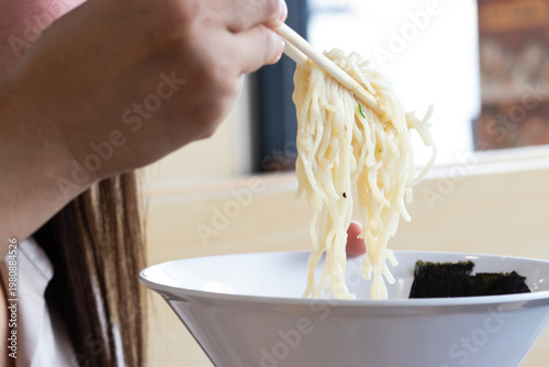Close-up of a person using chopsticks to lift ramen noodles from a white bowl, showcasing the texture and steam in a lifestyle dining scene.
