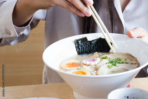 Close-up of a person using chopsticks to eat a bowl of Japanese ramen with chashu pork, soft boiled eggs, and nori seaweed on a wooden table.