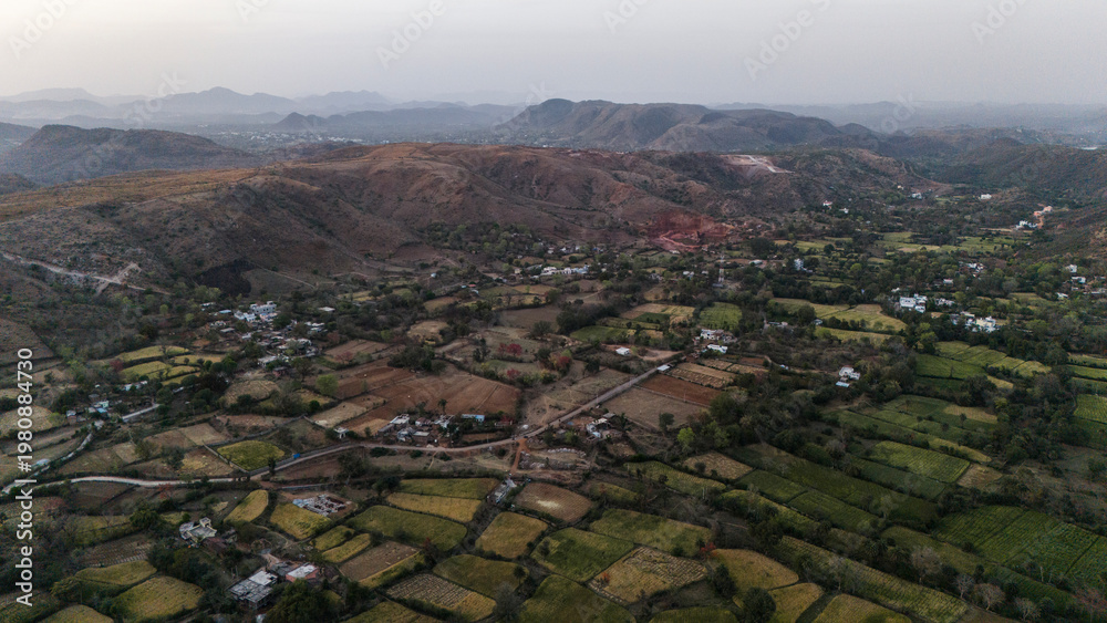 Obraz premium Aerial view of the Aravalli hills near Udaipur, Rajasthan at sunset, showing winding roads, patchwork agricultural fields, native trees, and scattered village houses across rugged terrain.
