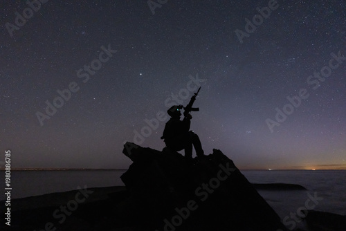 Bild auf Leinwand Silhouette of a lone tactical operator with night vision sitting on a rock near the ocean, holding a rifle under a vast starry sky