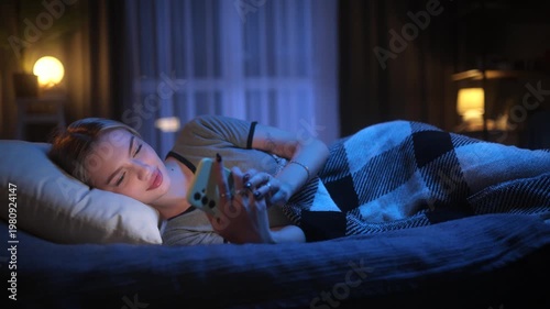 Happy young woman lying in bed and texting on smartphone late at night