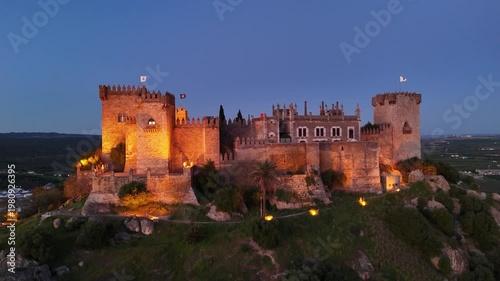 Aerial 4K view of the illuminated Almodovar Castle at night in Cordoba, Spain. Medieval fortress with exterior lights on and scenic night landscape.