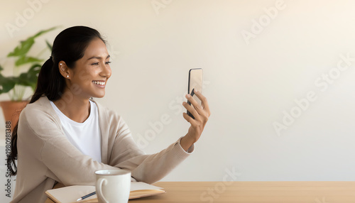 A woman smiles while video calling on her smartphone, sitting at a table with a notebook and coffee cup nearby.