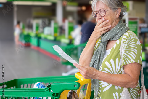 Worried senior woman standing in a supermarket checkout area, holding and reviewing her shopping receipt beside a grocery cart, expressing concern about household expenses and rising food prices