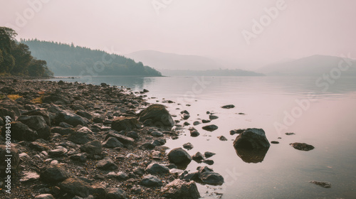 Rocky lakeshore with tranquil water reflecting soft sky, distant hills fading into atmospheric light