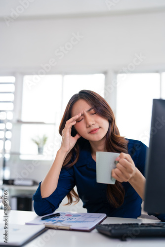Stressed business woman rubs her temple while holding a coffee cup and working on a laptop in office, fatigue, frustration and  stress.