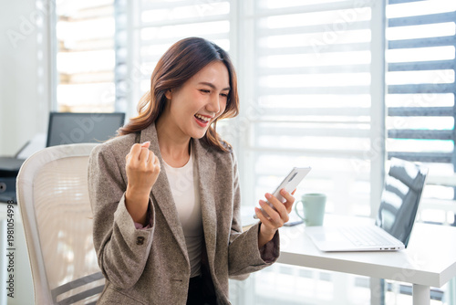 Asian business woman getting bonus reward, good news on telephone mobile phone at work in office. Happy, smiling ,winner, triumph, yes gesture.