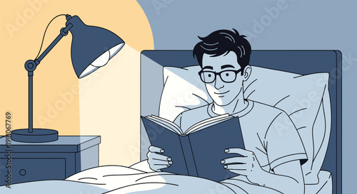 Young man lying in bed and reading an interesting book under the light of a desk lamp in a dark bedroom before sleeping.