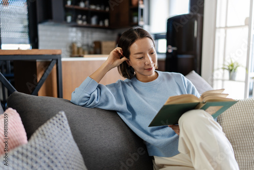 Asian woman relaxing on a sofa reading a book at home. Cozy lifestyle scene showing calm, focus and quiet leisure time in a modern living room interior
