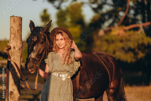 Tableau sur toile A young woman with a horse stands near a fence in autumn at sunset