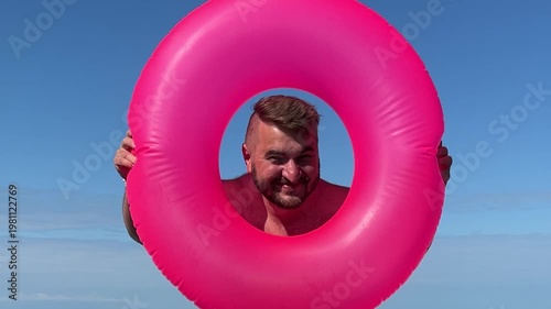 Horizontal travel video. Young adult white fat man with beard, stubble, big belly stands on ocean shore or Black Sea pebble beach looks out from pink swim ring for swimming at camera. Tourism, trip