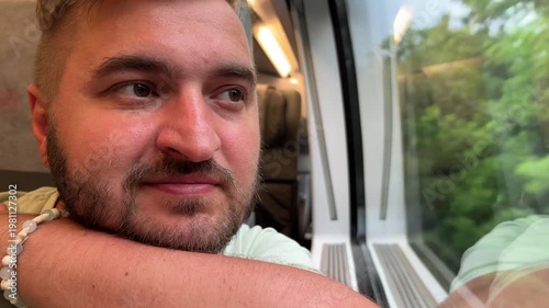 Travel 4K video close up. 60 fps. Young adult fat white man with beard, green T-shirt sits on seat, smiles and looks out of large train window. Concept of tourism, road trip, rest, relax, arrival, way