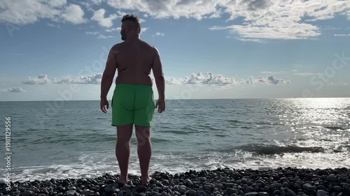 Horizontal 4K video. Back view. Young adult tanned fat white man with beard, stubble, big belly, green shorts on Black sea pebble beach looks around at sun day. Blue sky, wave. Tourism, travel concept
