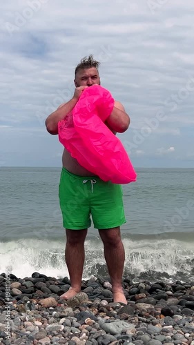 Vertical day video. Young adult fat white man with big belly, green shorts blows pink swim ring for swimming on pebble beach coast. Blue sky, sea wave. Summer vacation, rest, trip, travel concept