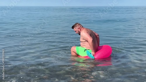 Travel video. 60 fps. Mature white fat man with beard, green shorts, big belly, blue slates tries to sit in pink magenta swim ring in sea and falls. Shore, pebble beach. Tourism. Blue sky. Ocean wave