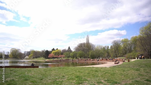 Wide shot of people relaxing on wooden amphitheater seating by a lake in a public park under a cloudy sky