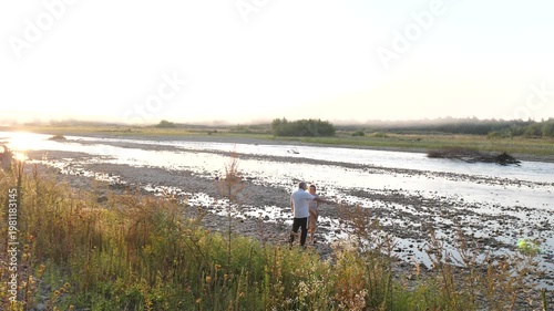 A man and a woman stand on a rocky riverbank watching the water under soft evening light